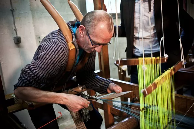 Fabrication d'une frange destinée aux canapés des salons ovales du théâtre. Actionnant les fils de soie tendus à la lève ou à la baisse par le biais de pédales, le tisseur assure l'entrecroisement nécessaire à la tenue des guipures qui forment les franges. Celles-ci sont calibrées grâce à une planchette qui en détermine la longueur. Le tisseur réalise 10 à 12 mètres par jour pour le modèle demandé. Il conviendra ensuite de retordre les brins de la frange, un à un, grâce à un moulinet et un crochet, pour leur donner leur régularité et une longueur égale.